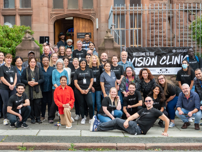 A large group of around 40 people stood outside a brick building, there is a banner which reads "Welcome to the Vision Clinic"