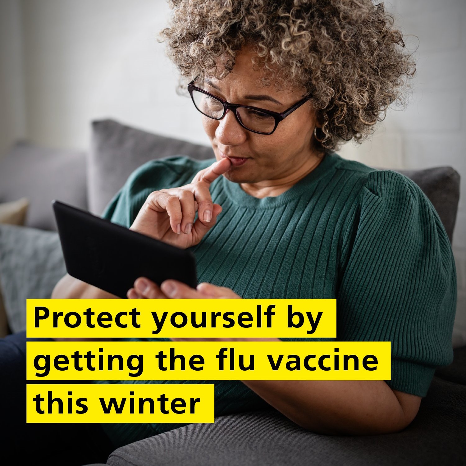 A woman with curly hair and glasses sits on a couch, reading a tablet. She looks focused. Text overlay: "Protect yourself by getting the flu vaccine this winter."