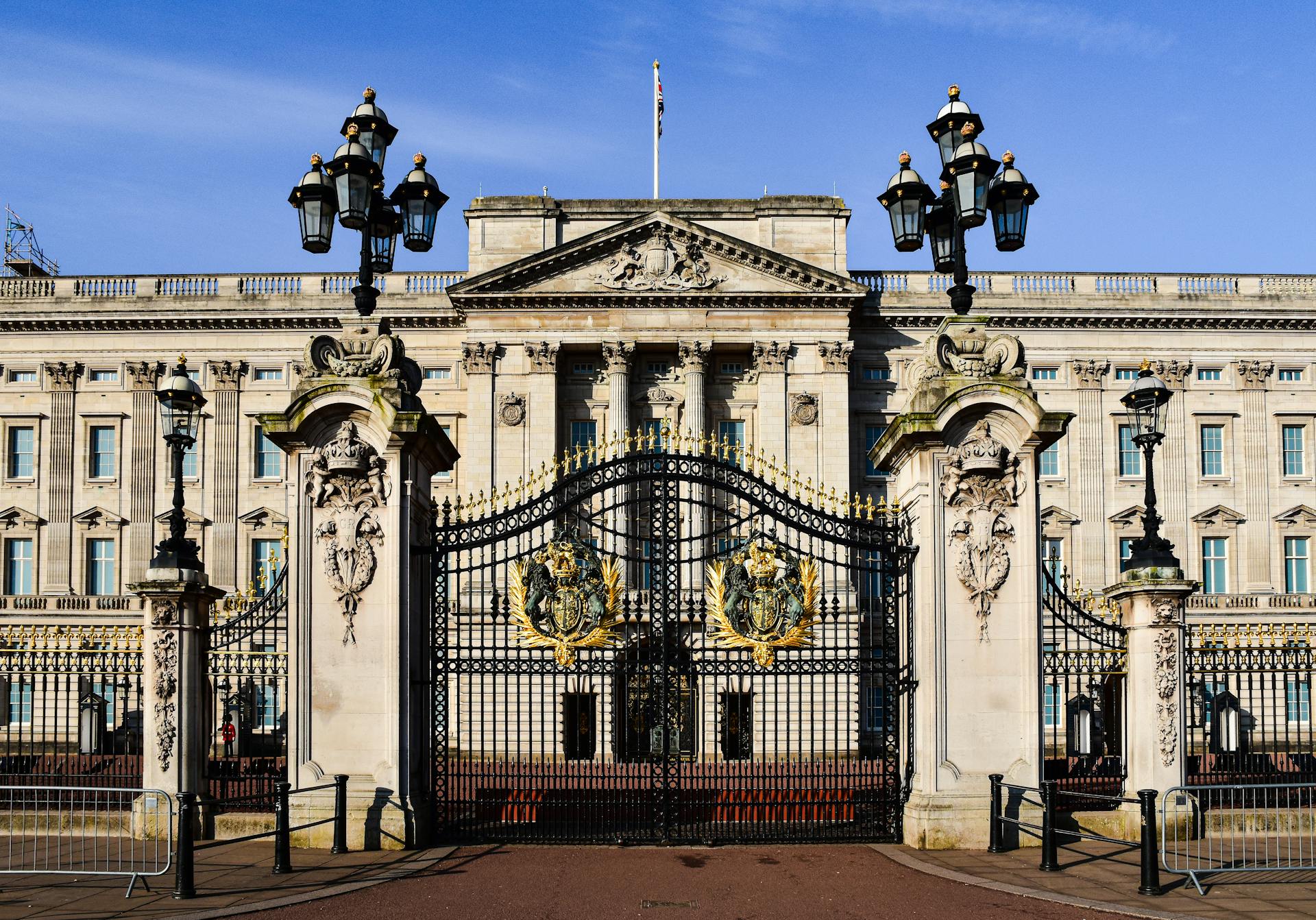 Elegant view of Buckingham Palace's grand facade and ornate black gates, adorned with golden crests, under a clear blue sky.