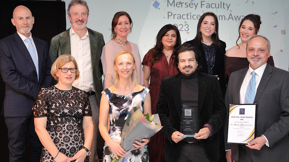 A group of ten people, smiling and posing at an awards ceremony. One person holds a trophy, another has a bouquet, and a third displays a framed certificate.